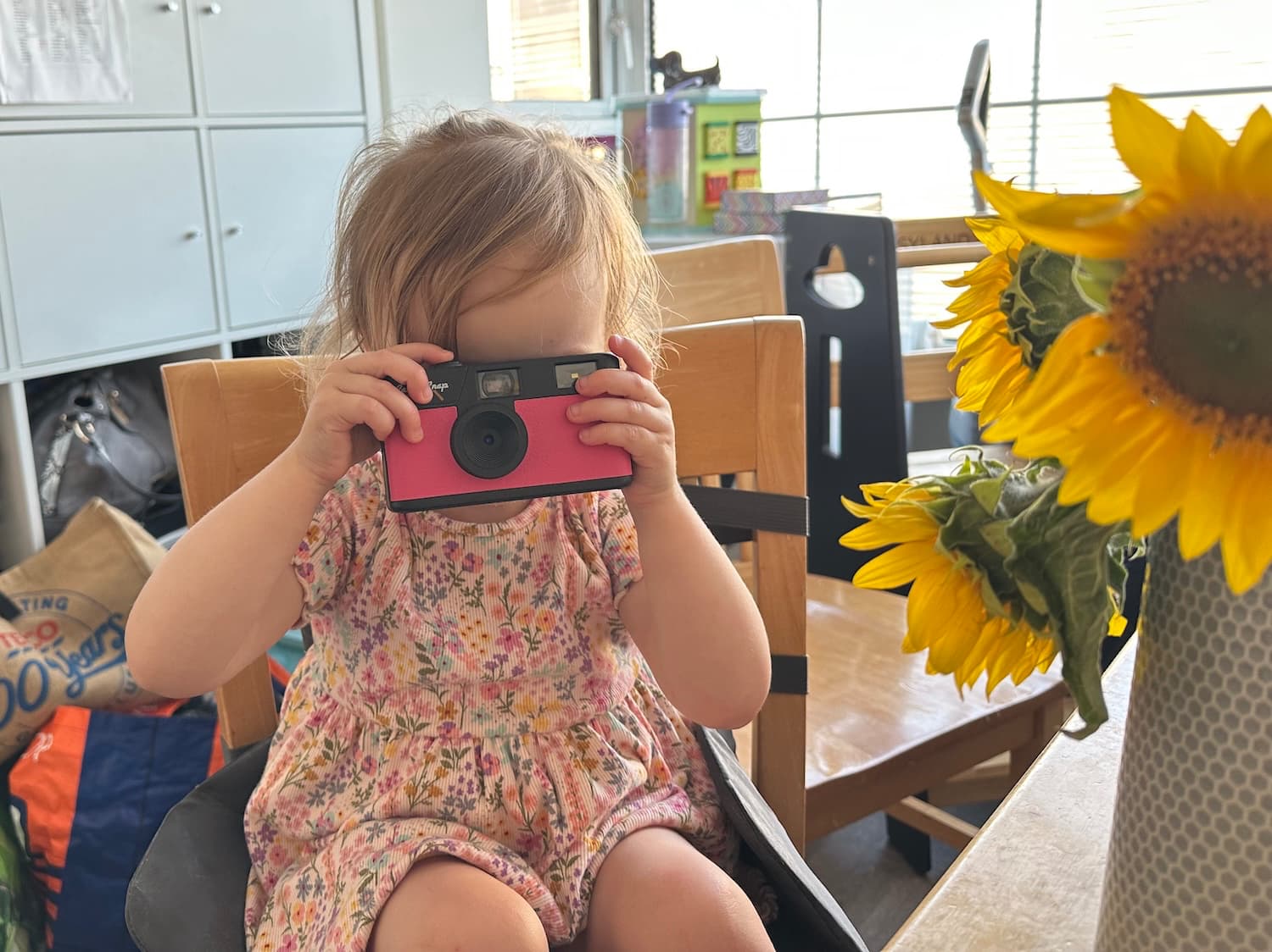 A child with blonde hair taking a photo with a pink camera