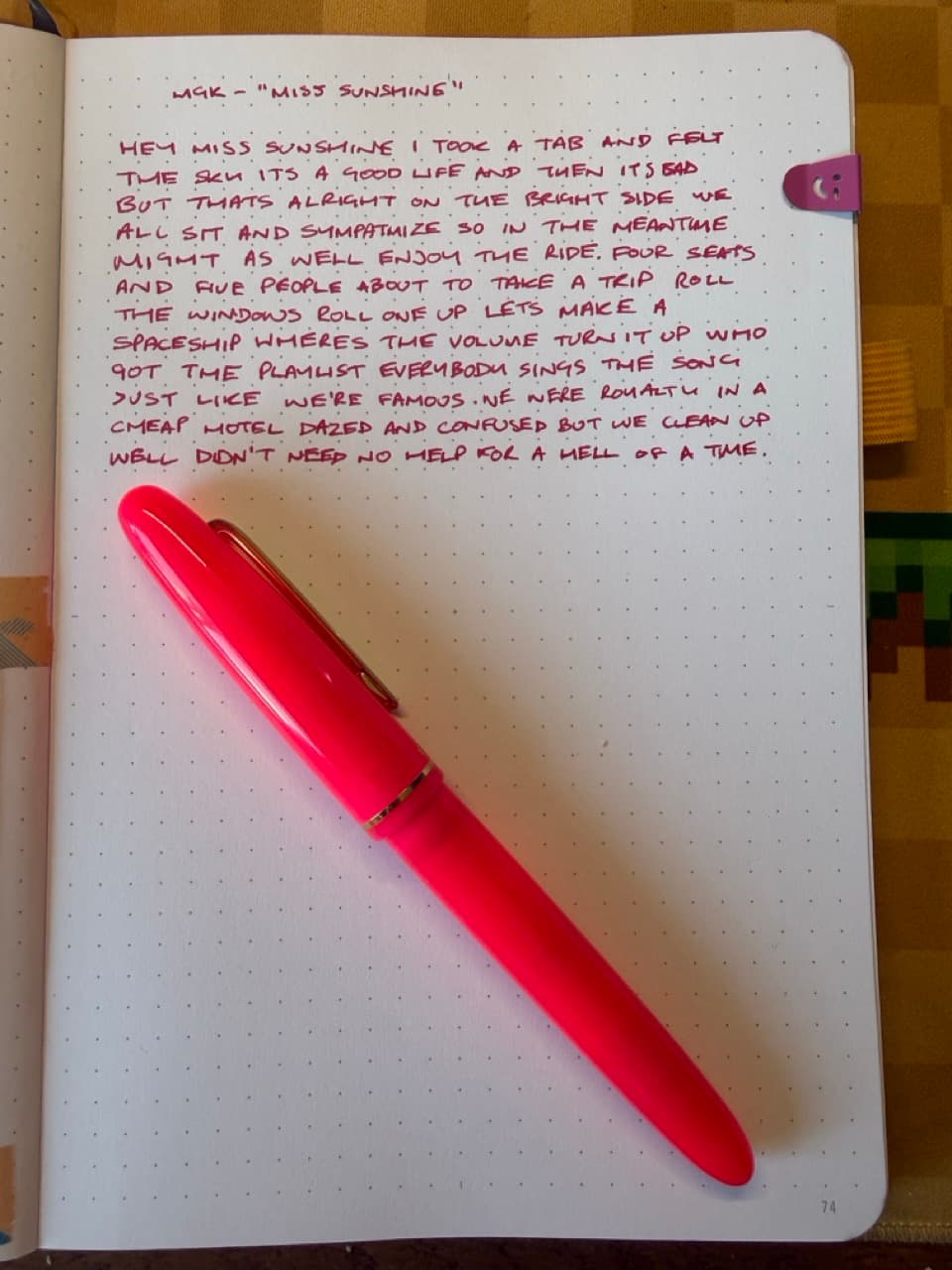 A page in a notebook with handwritten lyrics to MGKs Miss Sunshine song. A pink fountain pen is resting on the page below the writing. There's a smiley face paper clip on the right.
