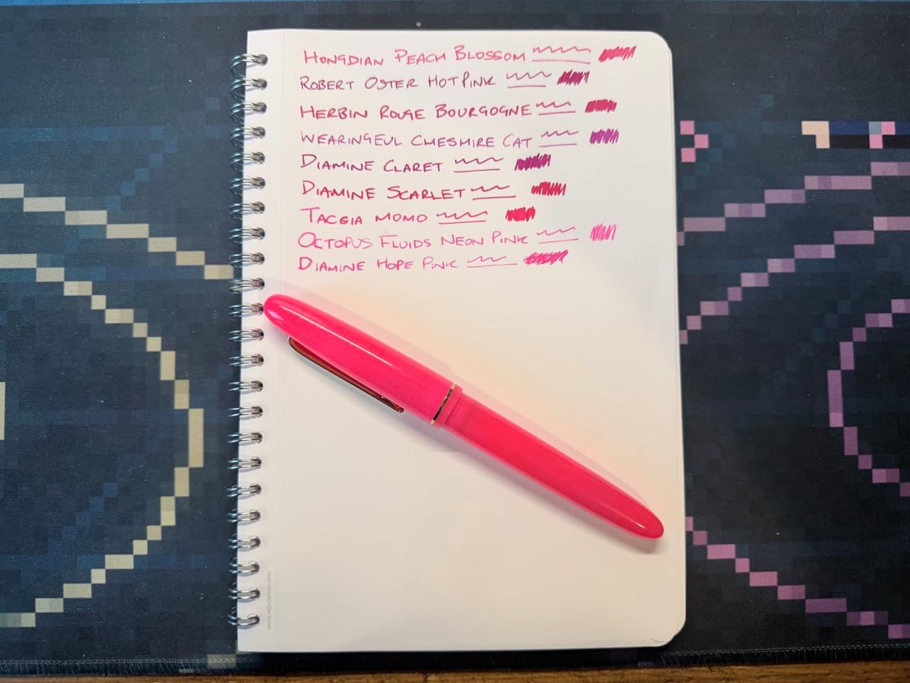 A notepad showing nine different ink samples all of various shades of pink and purple. Below is a bright pink fountain pen.