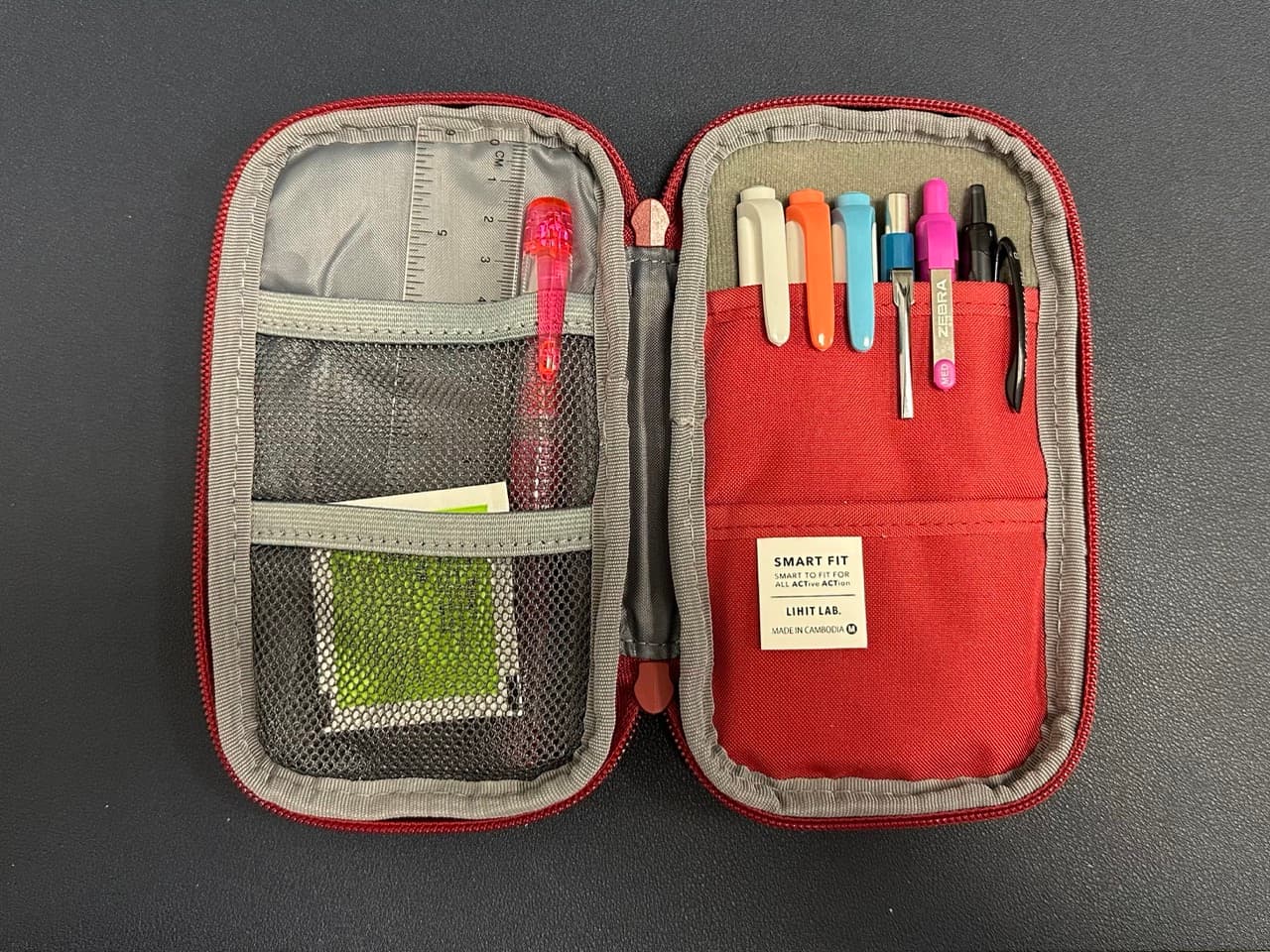 A red lihit labs pen case open on a desk with pens, pencils, and a ruler in it