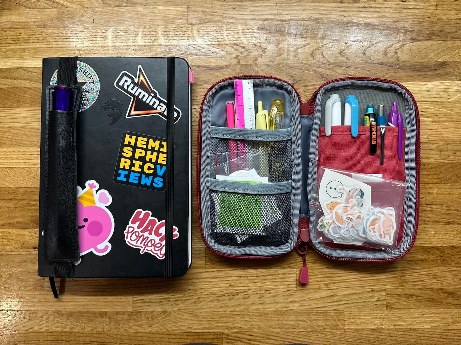 A notebook and pencil case on a wooden tabletop. The notebook has stickers on it and a pen attached in a leather holder.