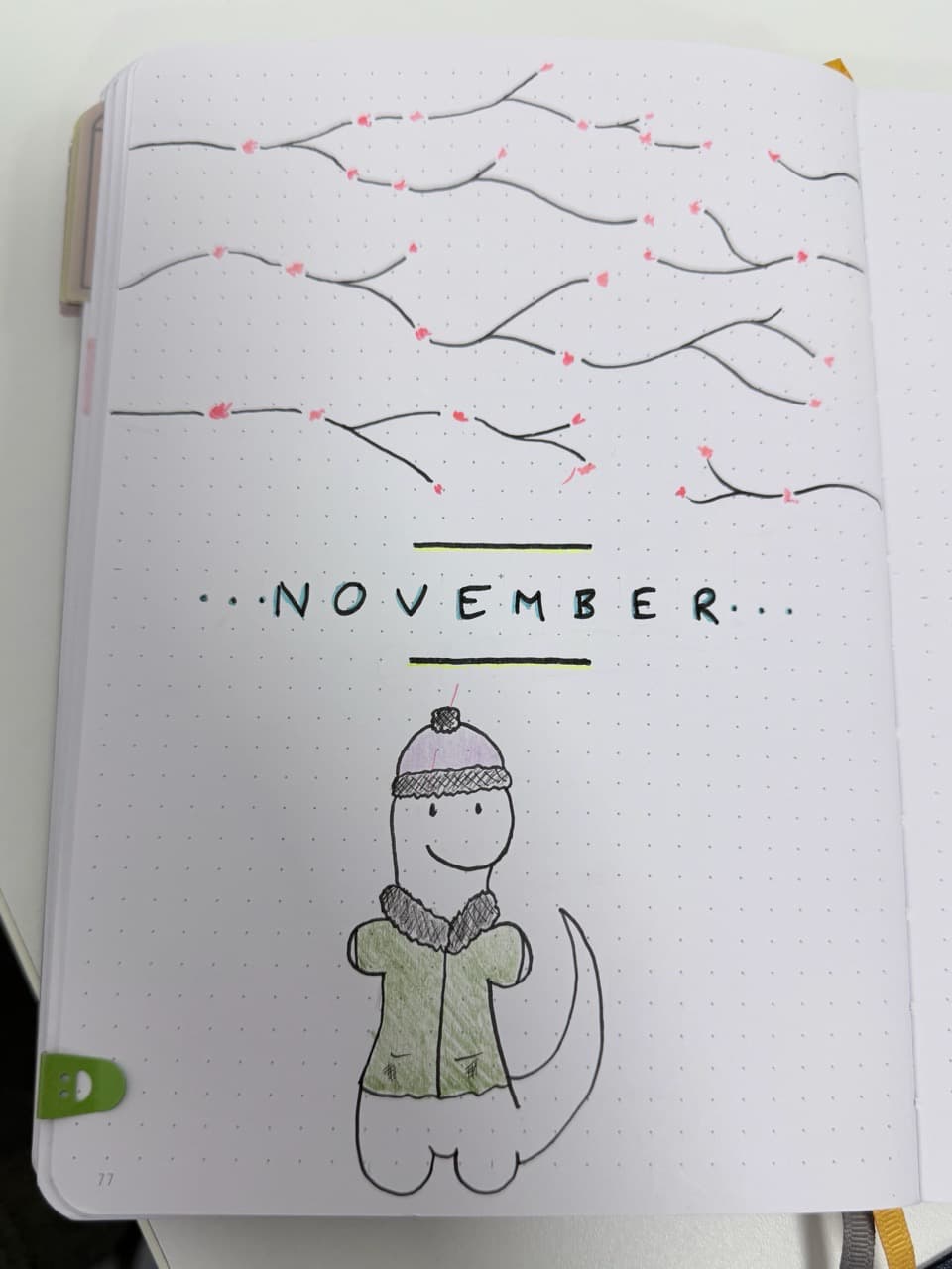 A dotgrid journal page. The top has tree branchs with pink flowers on it, then it says November in capitals in the middle. Below is a dinosaur in a wooly hat and coat.