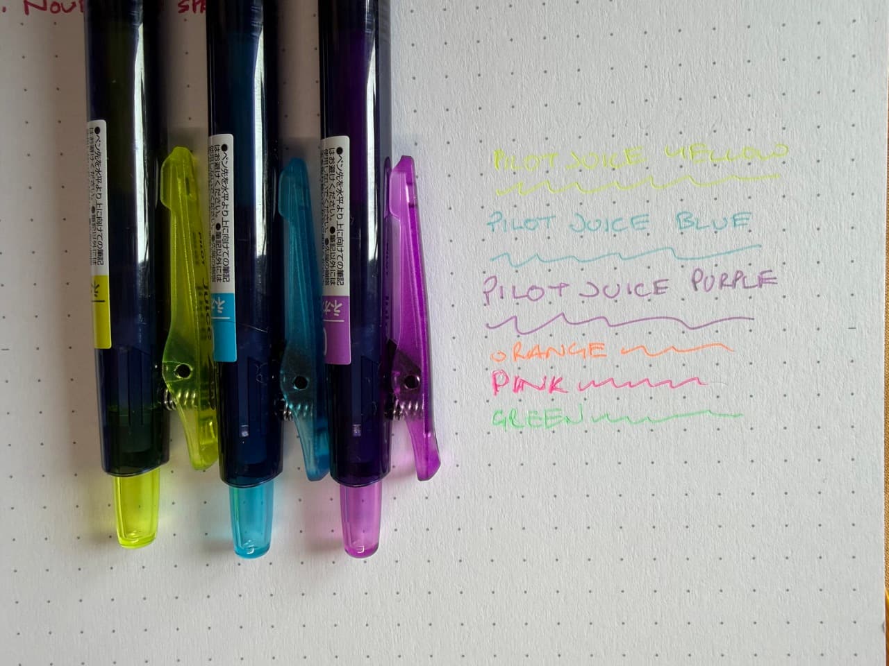 Three pens next to writing samples in yellow, blue, purple. Other samples are orange, pink, and green. The yellow is very hard to read