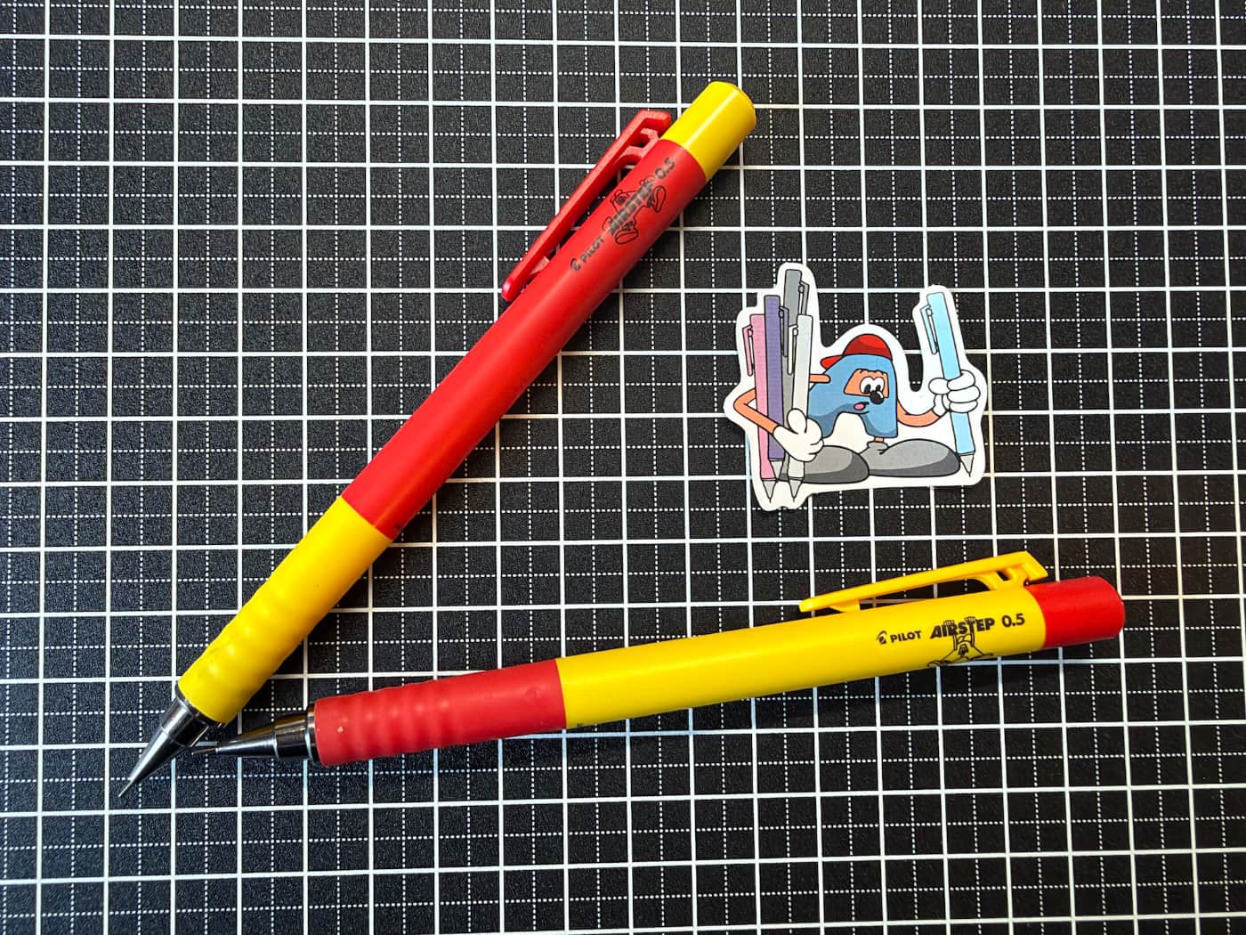Two red and yellow pencils on a black writing board with a sticker of a little pen character. He's called AirSteppy