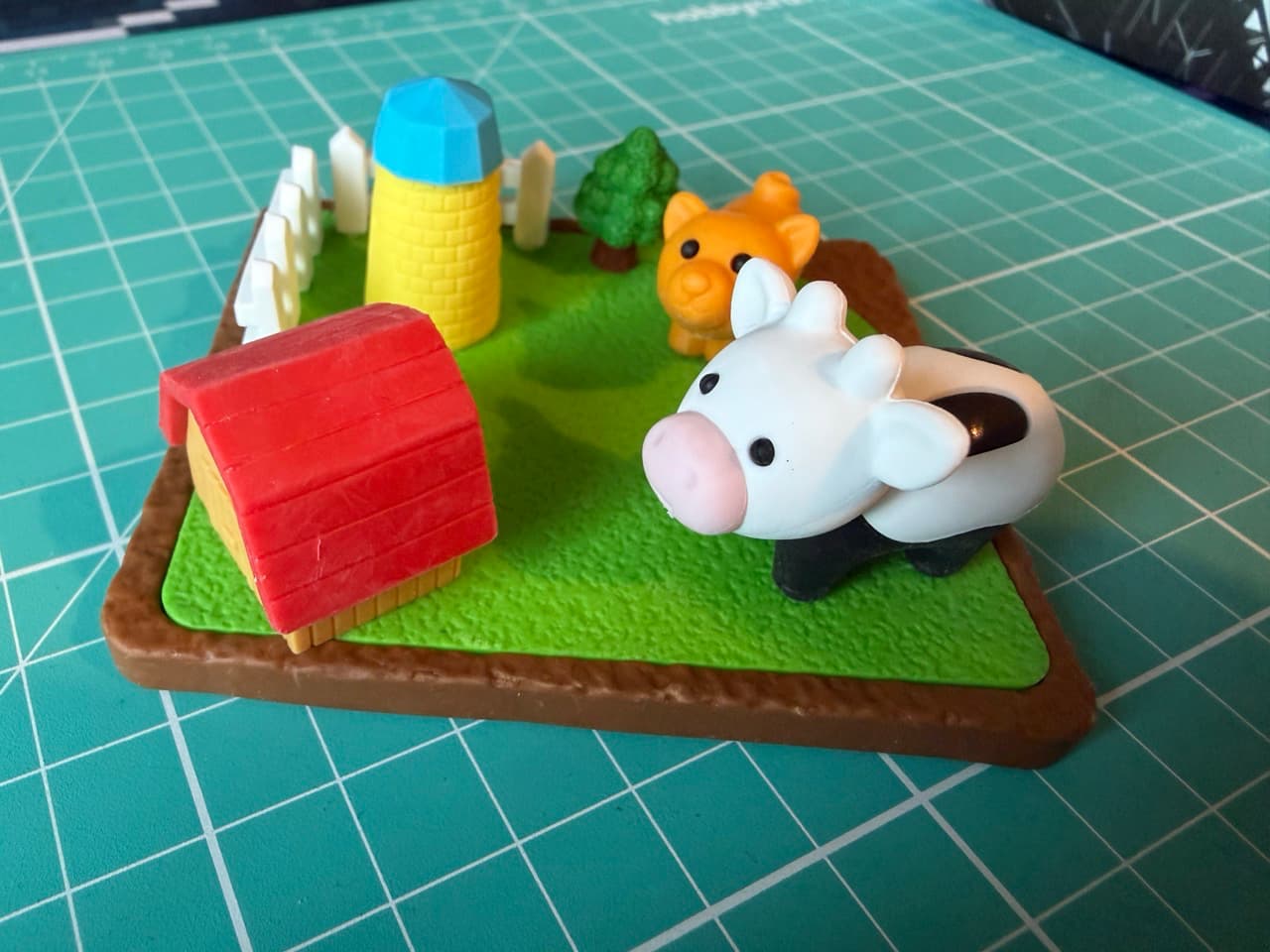 A small rubber farm with a cow, cat, and a barn on a green field with a white fence