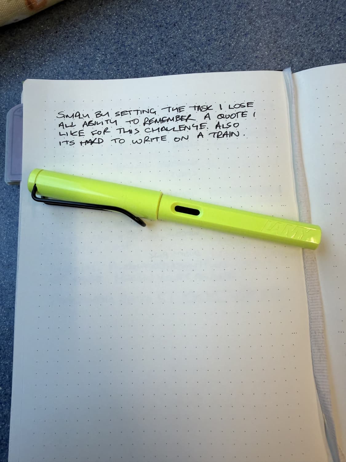 A notebook with a note written on it that says Simply by setting the task I lose all ability to remember a quote I like for this challenge. Also it's hard to write on a train. A neon pen is underneath