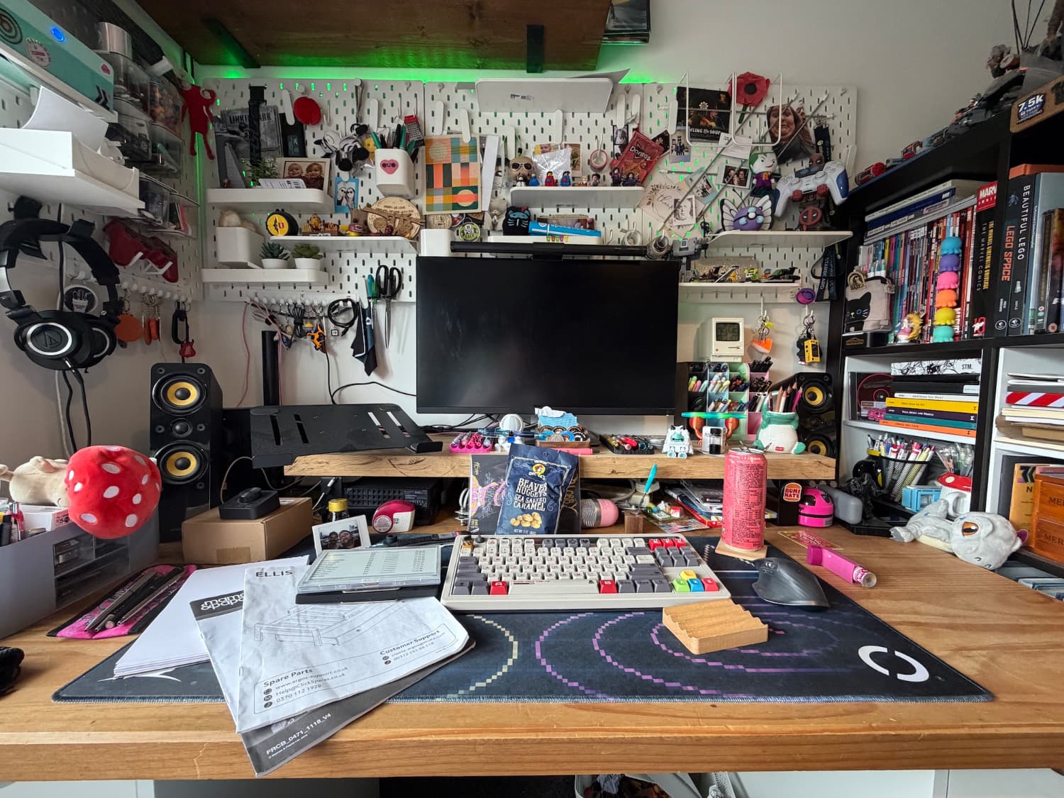 A desk with lots of stuff on it including pegboard behind it with photos and other nick nacks. The desktop has paperwork and other bits on it.