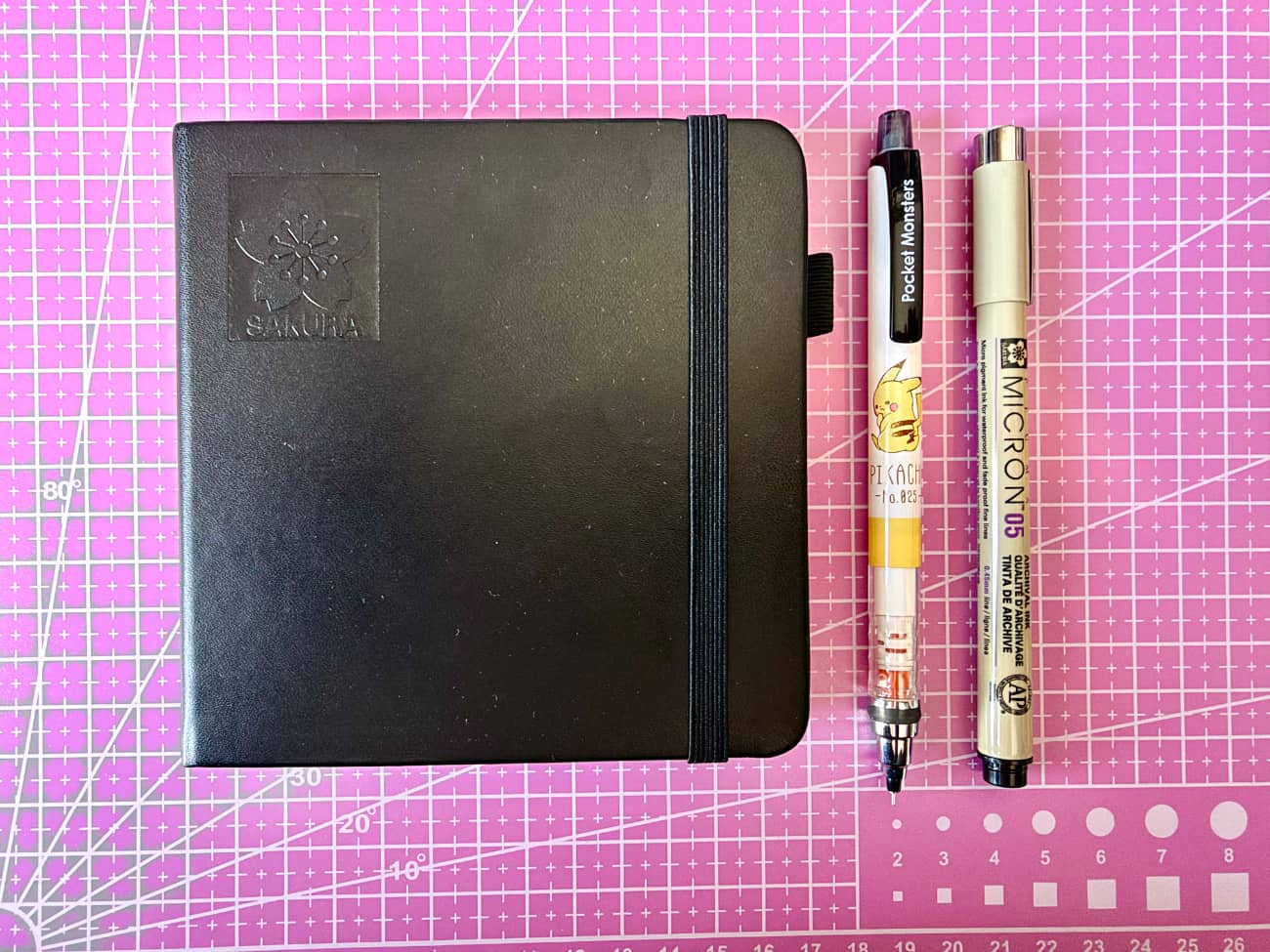 A pink cutting board with a black square notebook on it. Next to it is a mechanical pencil with Pikachu on it and a Sakura micron fineliner