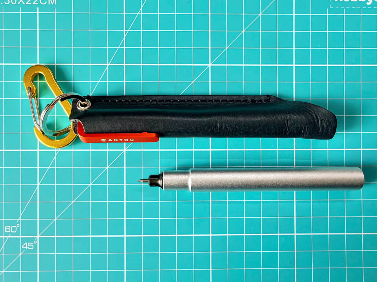 A leather sleeve and pen on a green cutting board. The leather sleeve has a carabiner attached to it