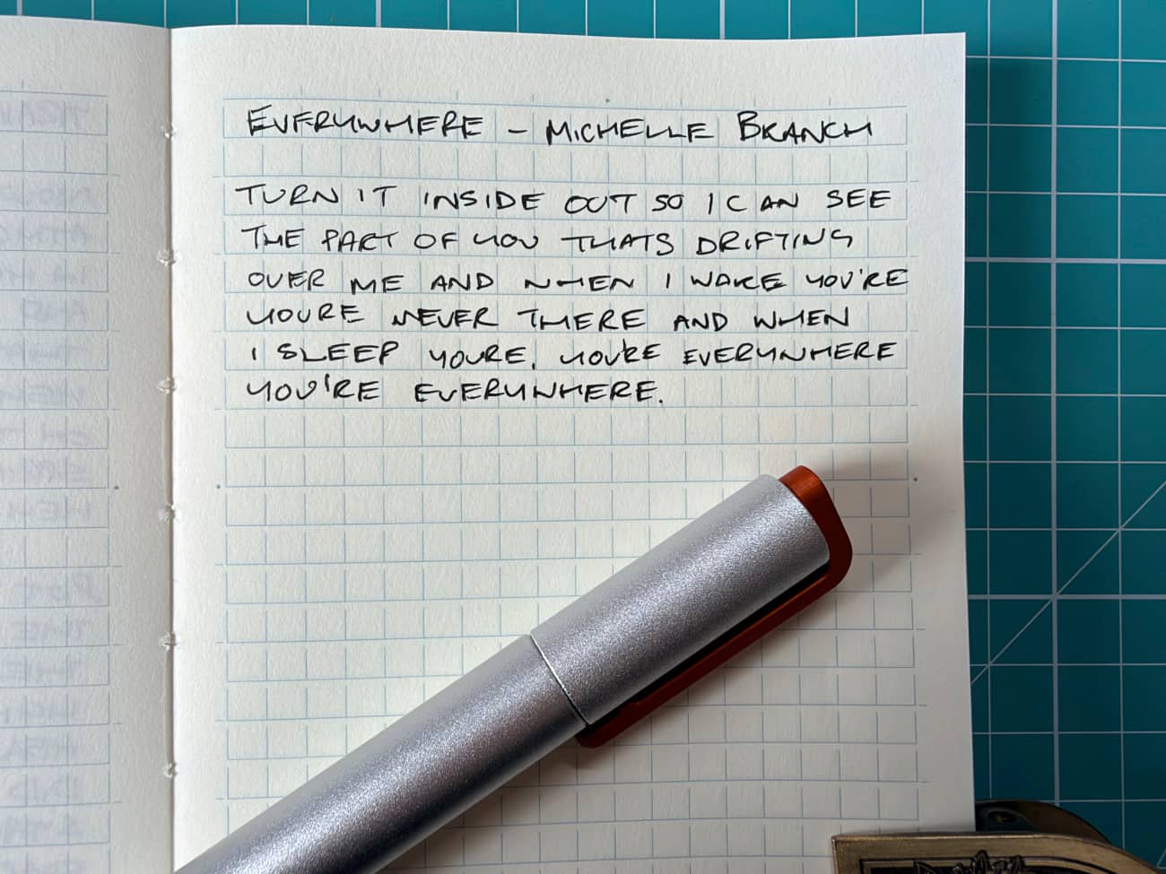 A notepad with a metal pen on top of it. The writing is the first verse to Michelle Branch Everywhere, noted at the top of the page
